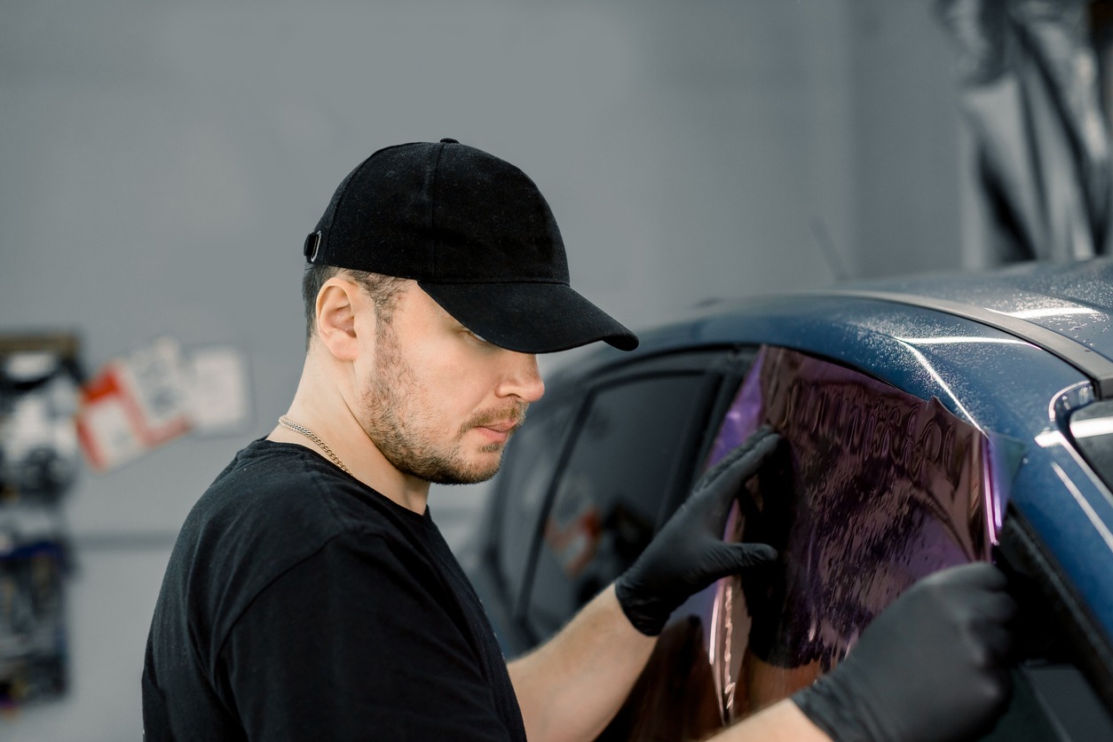 Professional car service worker wearing black cap and t-shirt, tinting a car window with tinted foil or film in auto workshop. Tinting of car windows