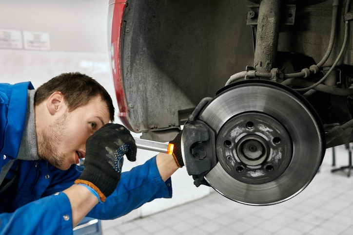 Male mechanic with open mouth wearing black gloves and blue uniform, holding flashlight and carefully examining tyres or brake pads of lifted car at auto workshop.Car service and technician concept