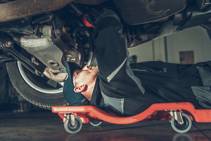 Caucasian Car Mechanic on a Creeper Performing Vehicle Repair Inside Authorized Auto Service Station.
