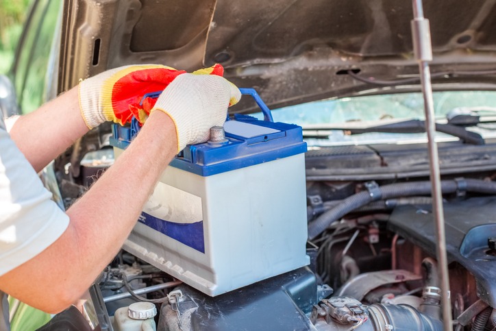  a car mechanic taking out a battery with a car battery carrier from under the hood of an auto
